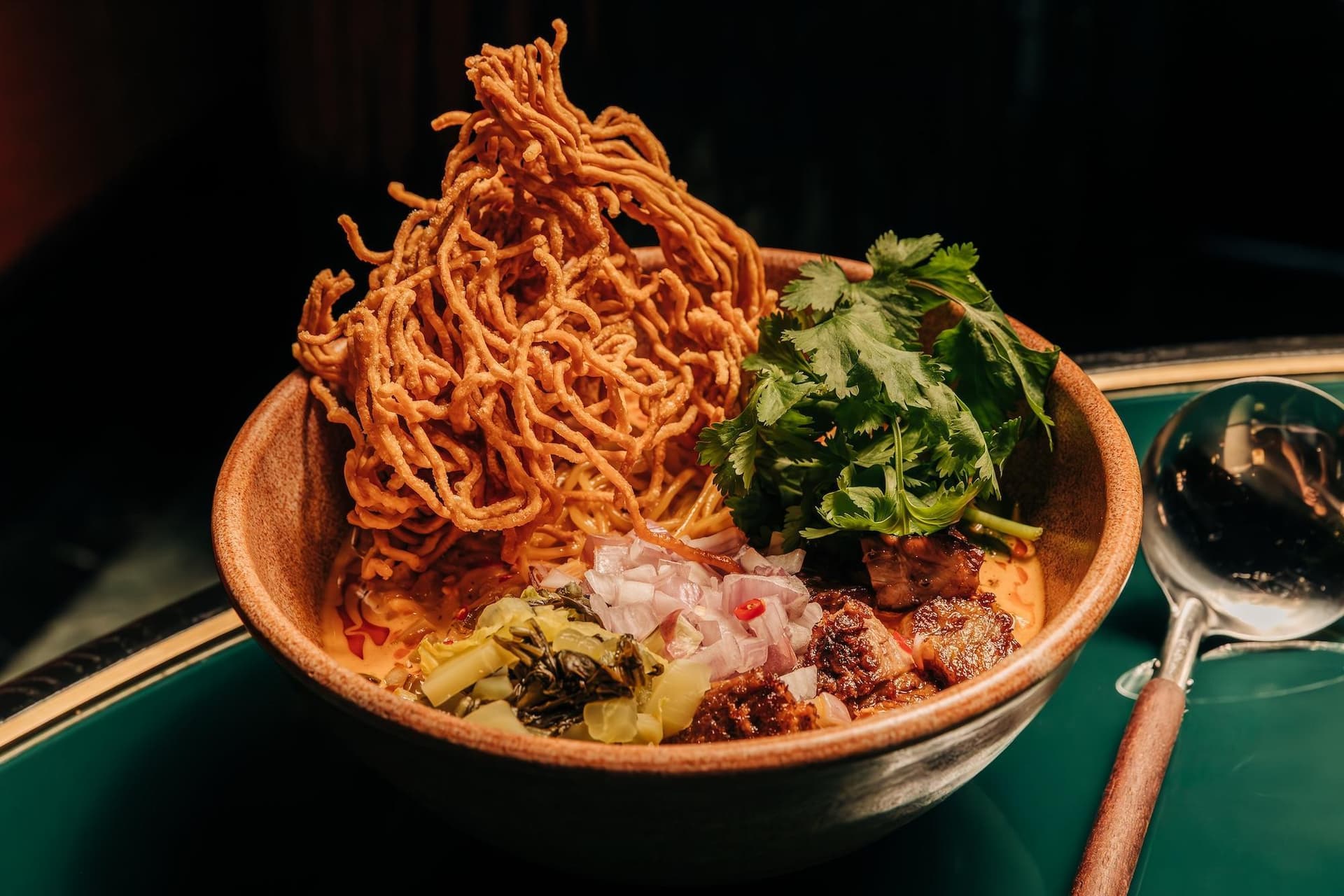 A bowl of Khao Soi, a Thai noodle dish, featuring crispy noodles on top, surrounded by fresh cilantro, chopped onions, and tender meat in a creamy curry broth.