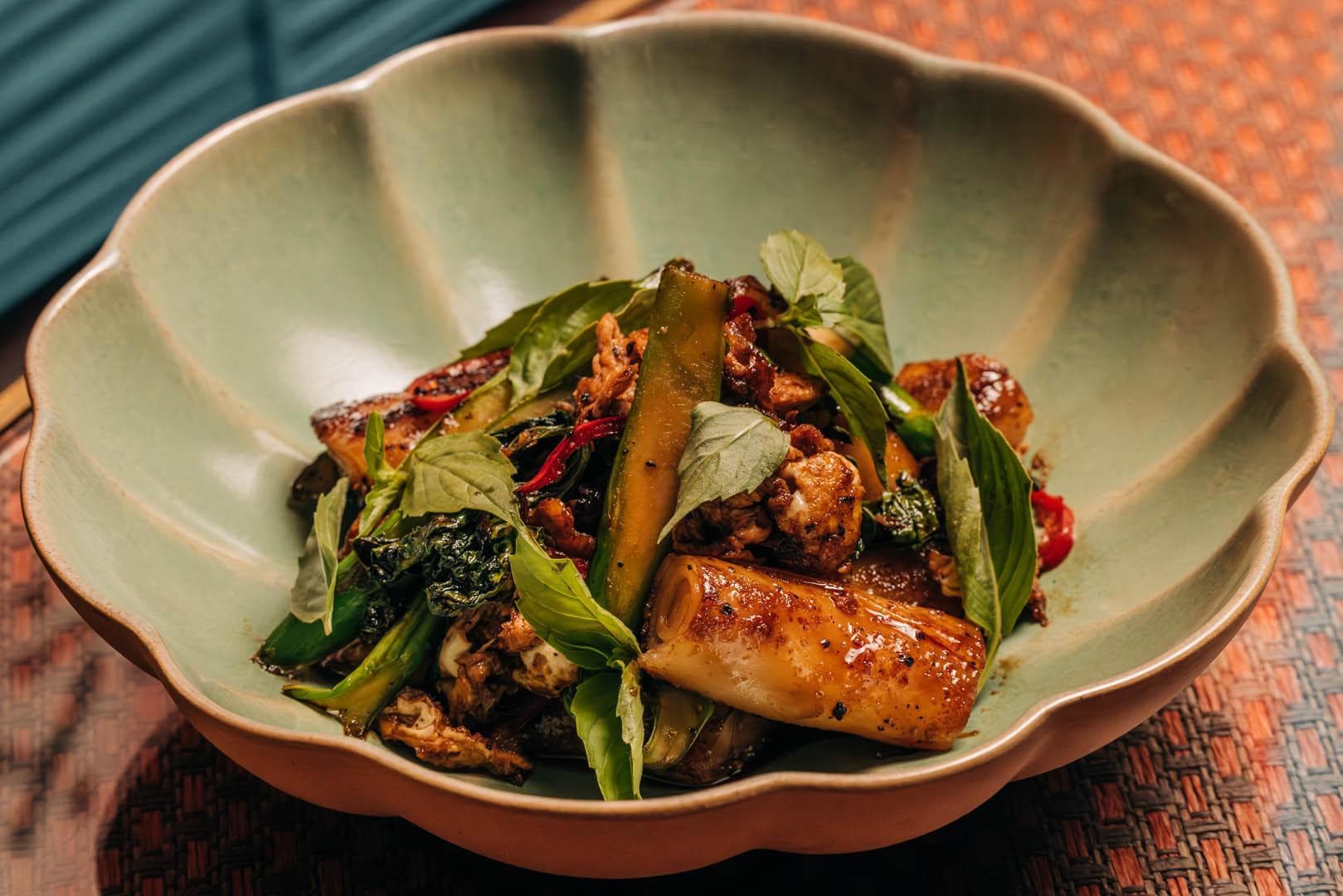A dish of stir-fried rolled rice noodles and vegetables garnished with fresh herbs, served in a scalloped, green ceramic bowl.