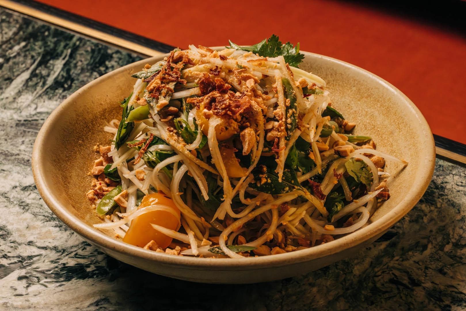A bowl of green papaya salad featuring shredded vegetables, herbs, and crushed peanuts, garnished with a cherry tomato on the side.