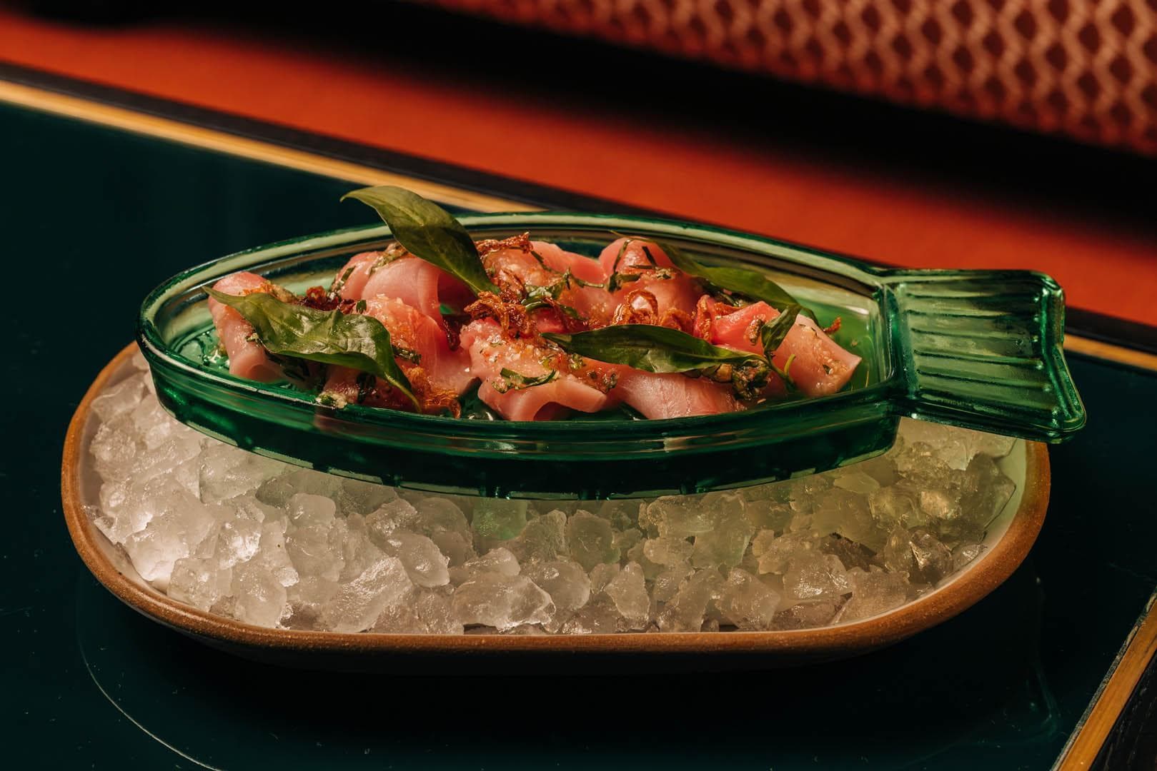 A glass dish filled with thinly sliced hamachi garnished with fresh green herbs, presented on a bed of crushed ice.