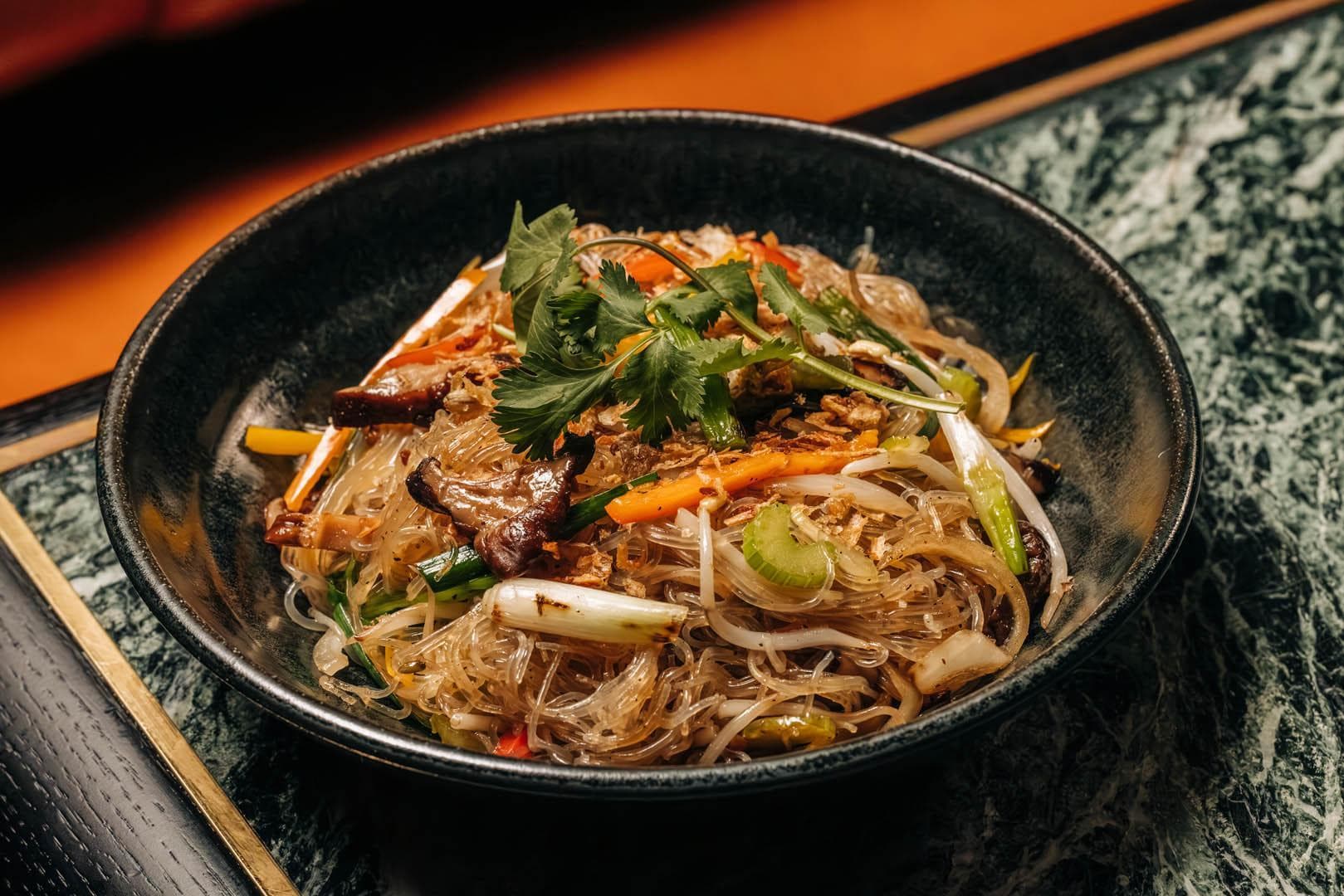 A bowl of stir-fried glass noodles with vegetables, garnished with cilantro and fried garlic, served in a dark dish on a textured surface.
