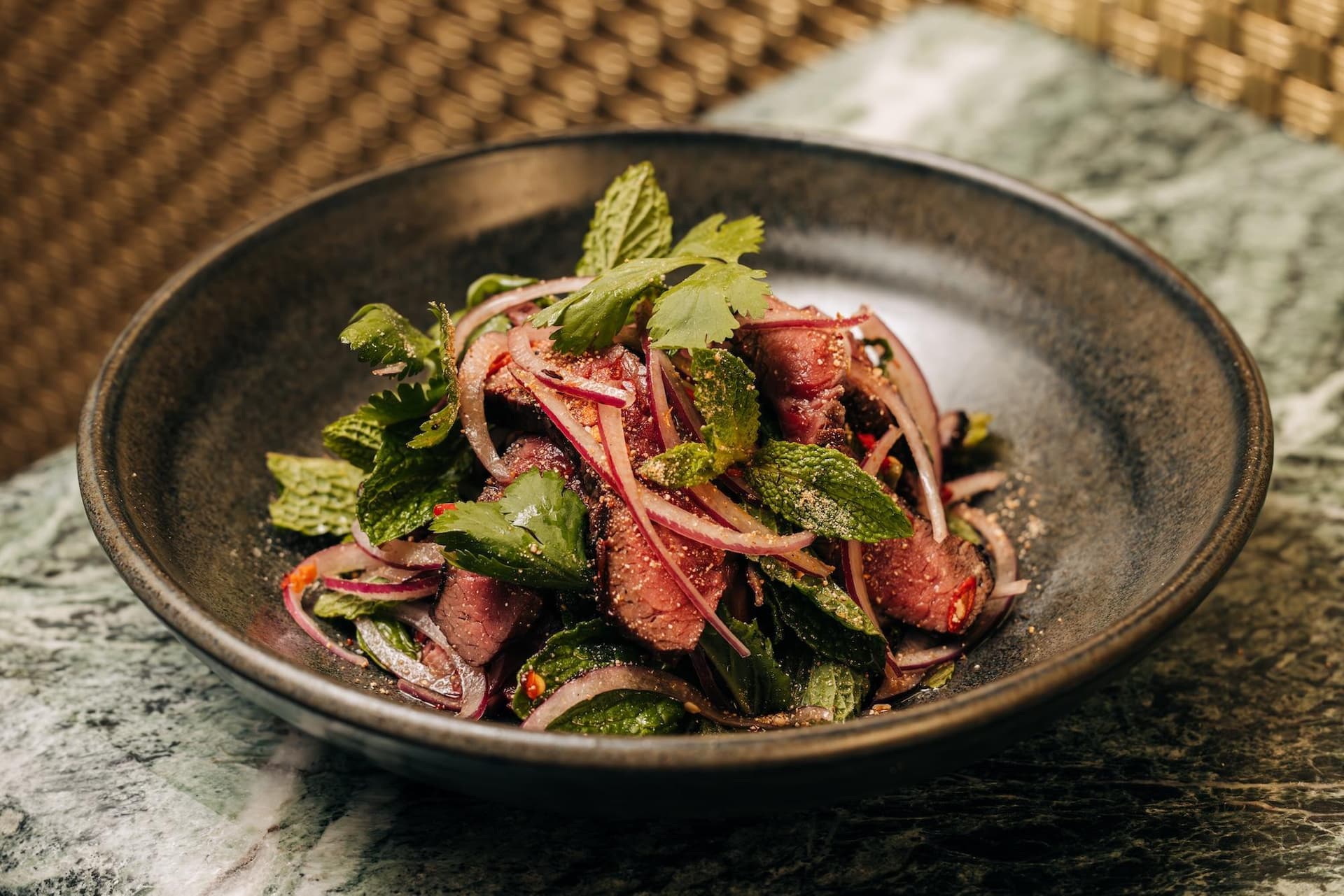 A bowl of Thai beef salad with sliced beef, red onion, mint leaves, cilantro, and green vegetables on a textured surface.
