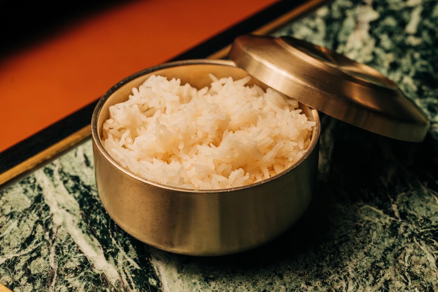 A brass container filled with cooked white rice, partially covered with a lid, placed on a decorative surface.