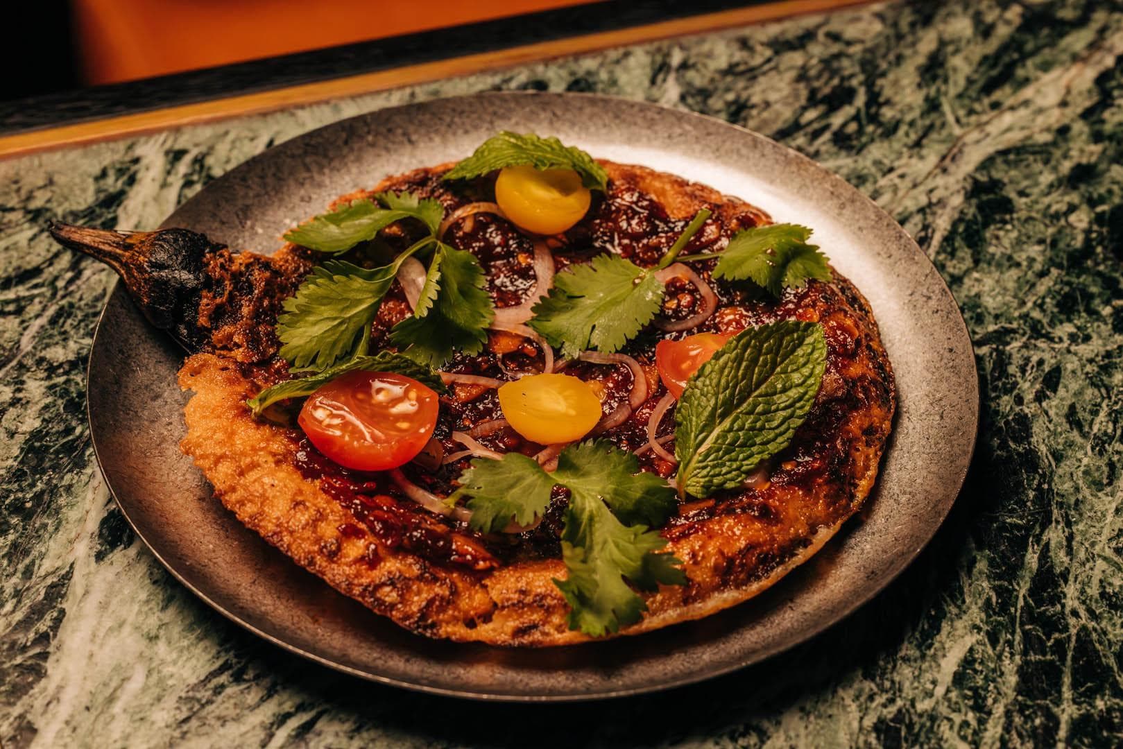 A delicious smashed eggplant topped with fresh coriander, sliced cherry tomatoes, and mint leaves on a textured, green marble table.