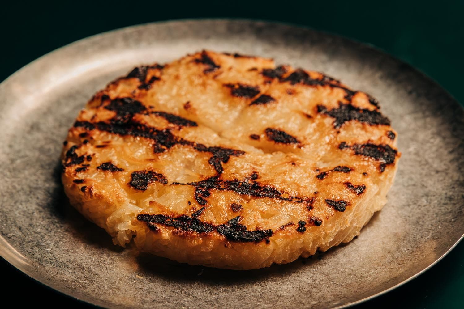 A grilled, round piece of grilled sticky rice with a slightly charred crust on a dark gray plate.