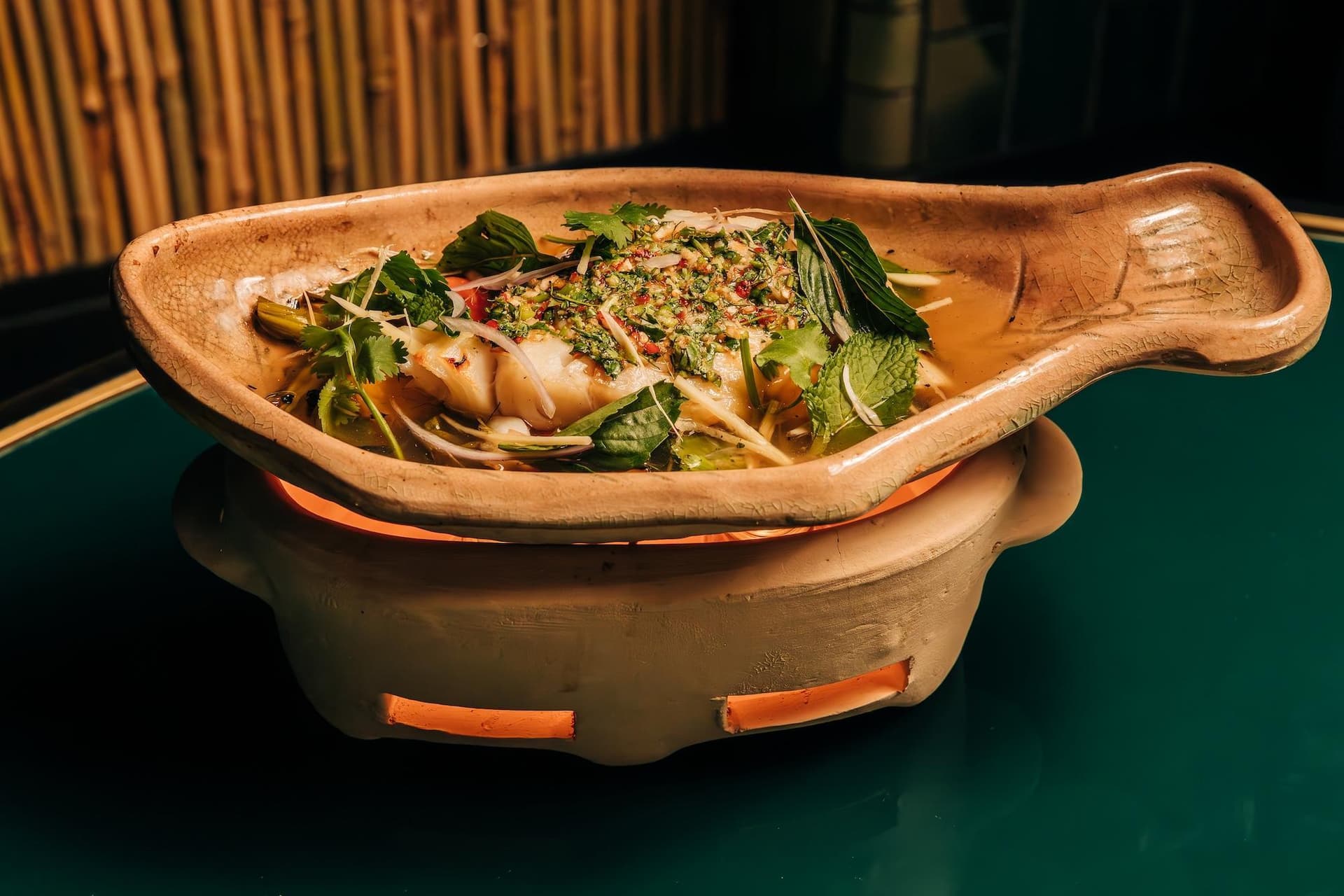 A dish of steamed fish topped with herbs and spices is presented in a clay pot, sitting on a small traditional stove.