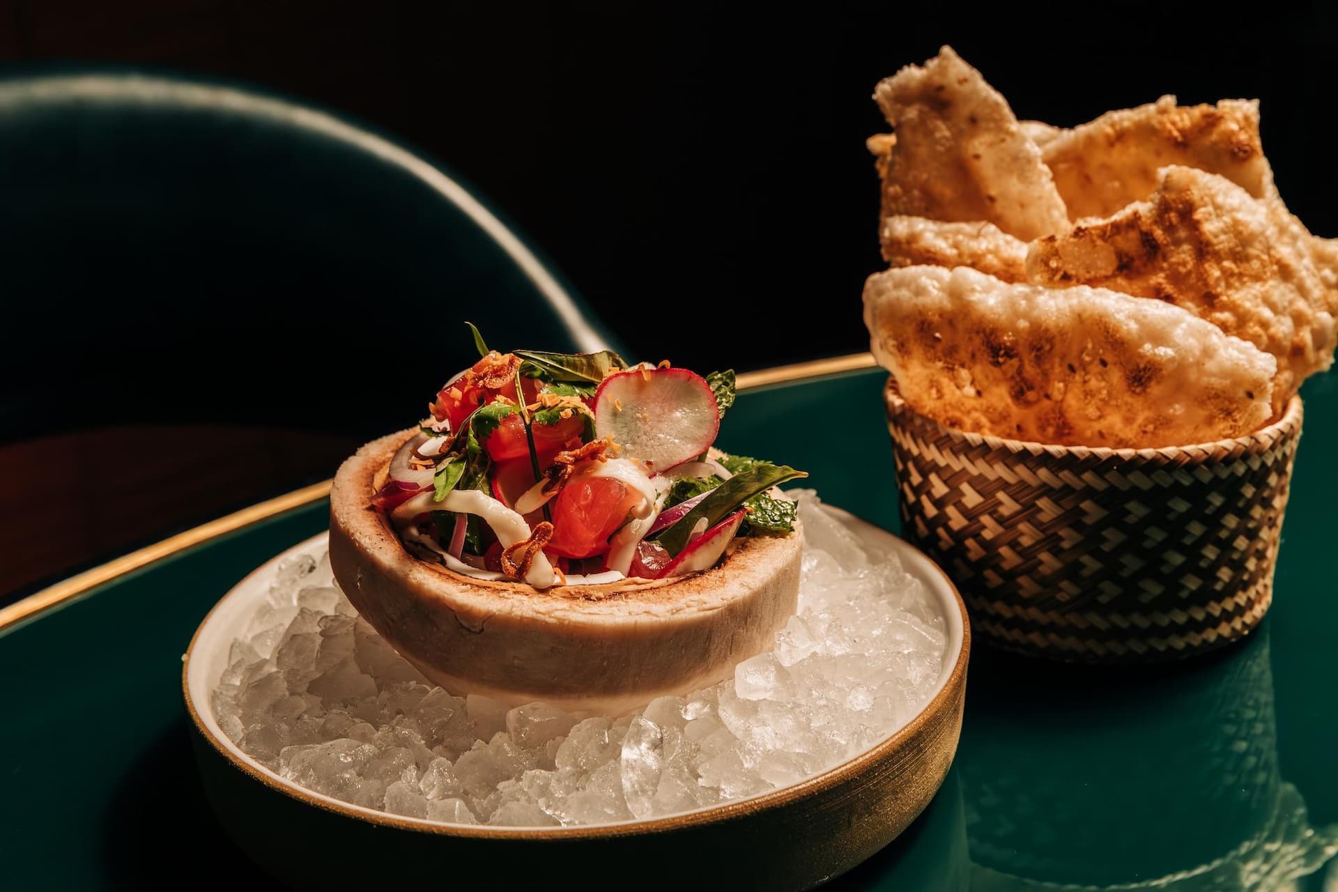 A dish of tuna tartare served in a halved coconut shell sits on a bed of ice, accompanied by a bowl of crispy chips.