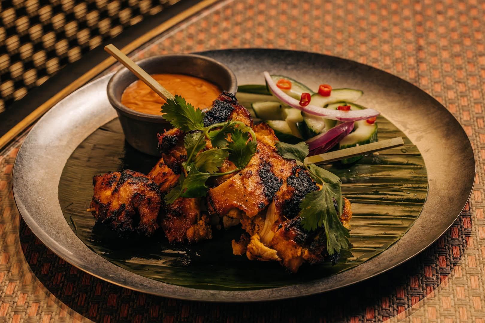 A platter featuring grilled chicken skewers garnished with fresh herbs. Accompanied by a small bowl of orange dipping sauce and a side of sliced cucumbers and onions with red chili slices. All served on a leaf-lined metal plate.