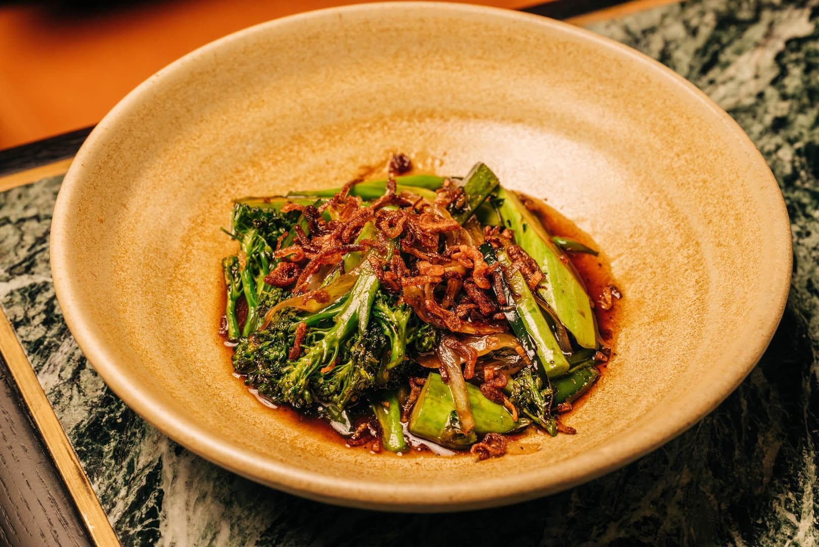 A dish featuring sautéed broccoli, garnished with crispy shallots and served in a brown bowl.