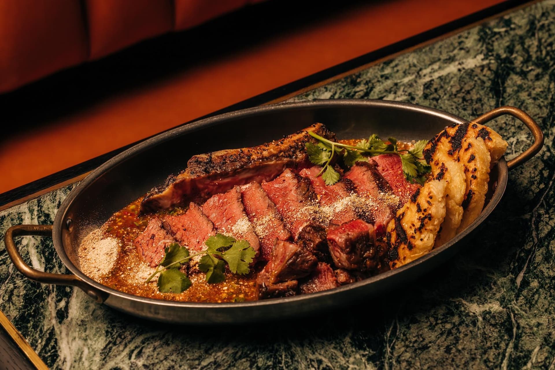 A dish of sliced grilled steak garnished with cilantro, served with grilled bread on a dark plate atop a marbled surface.