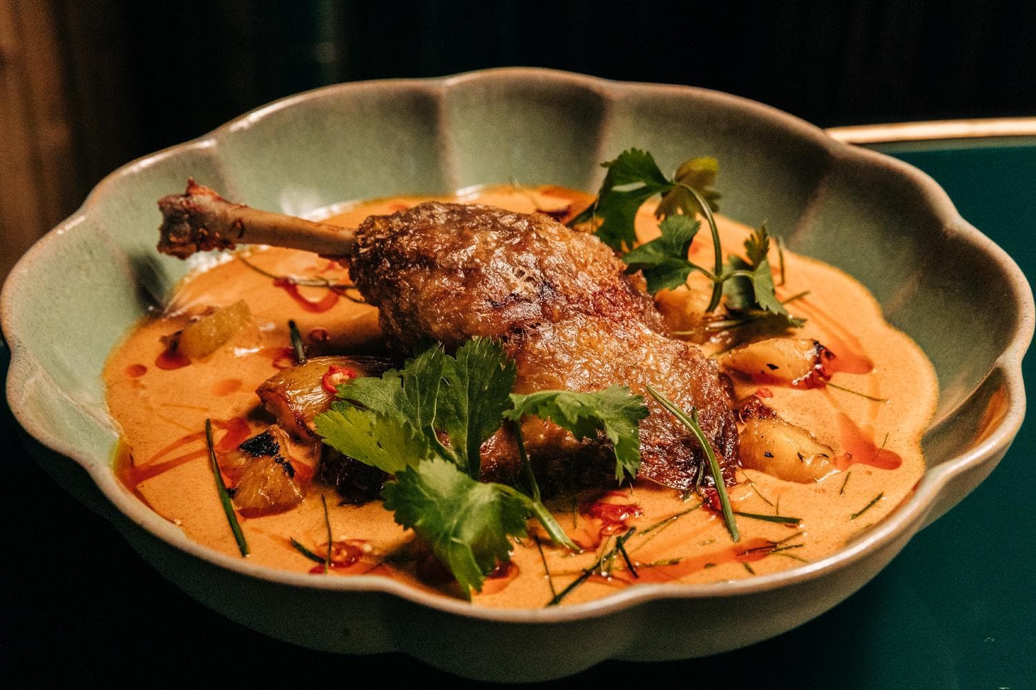 A dish featuring a cooked duck leg served on a bed of creamy curry sauce, garnished with fresh herbs and roasted vegetables, presented in a shallow, decorative bowl.