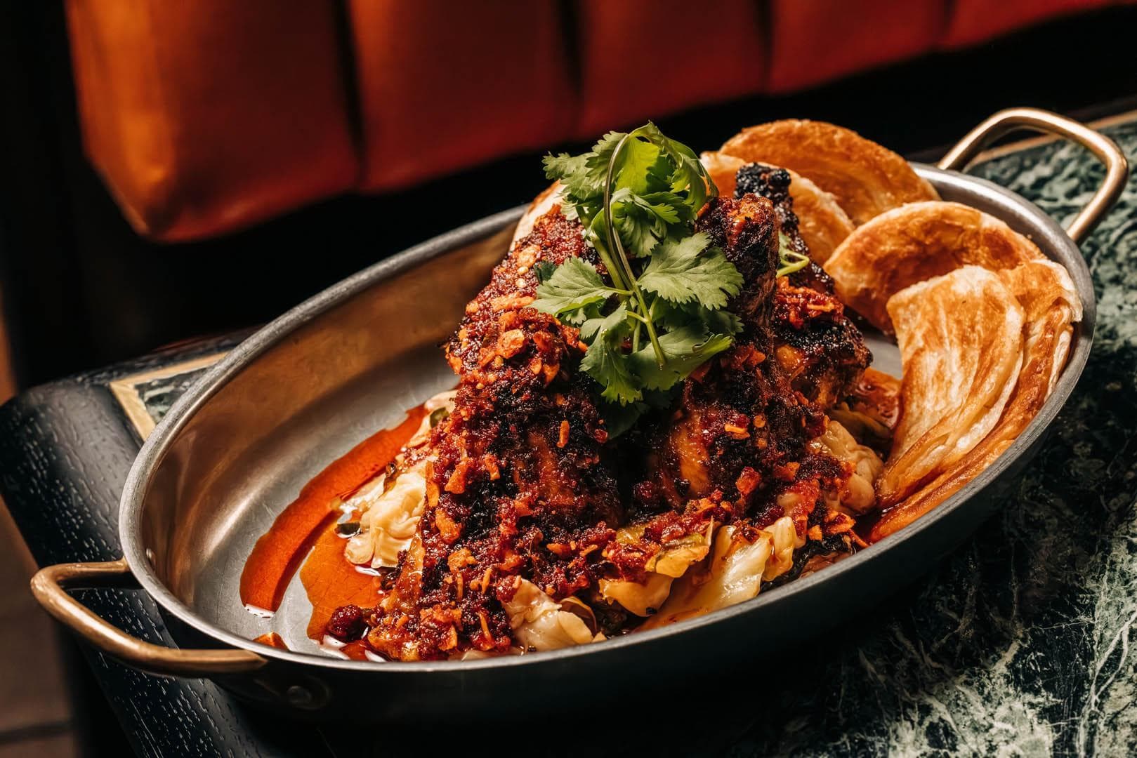 A dish of barbecue chicken topped with spicy red sauce and garnished with fresh cilantro, served in a metal pan. In the background, there is a blurred red cushioned seat.