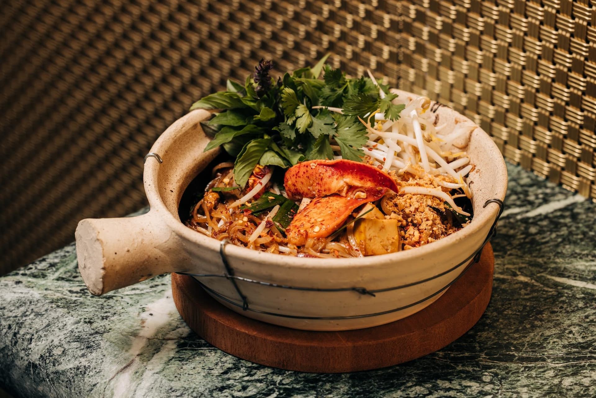 A ceramic bowl containing a noodle dish topped with herbs, bean sprouts, and lobster pieces. The bowl is placed on a wooden base, with a woven texture backdrop.