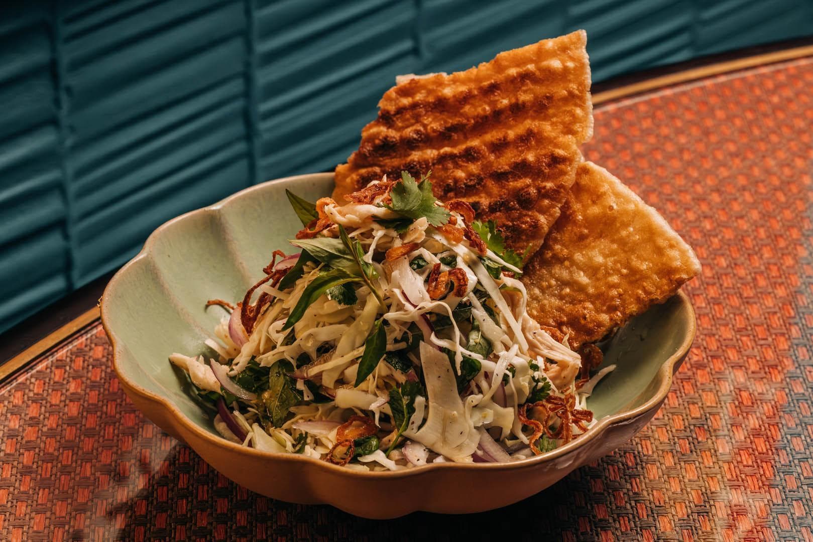 A plate of mixed vegetable and chicken salad with dressing, accompanied by crispy, toasted bread slices in a decorative bowl.