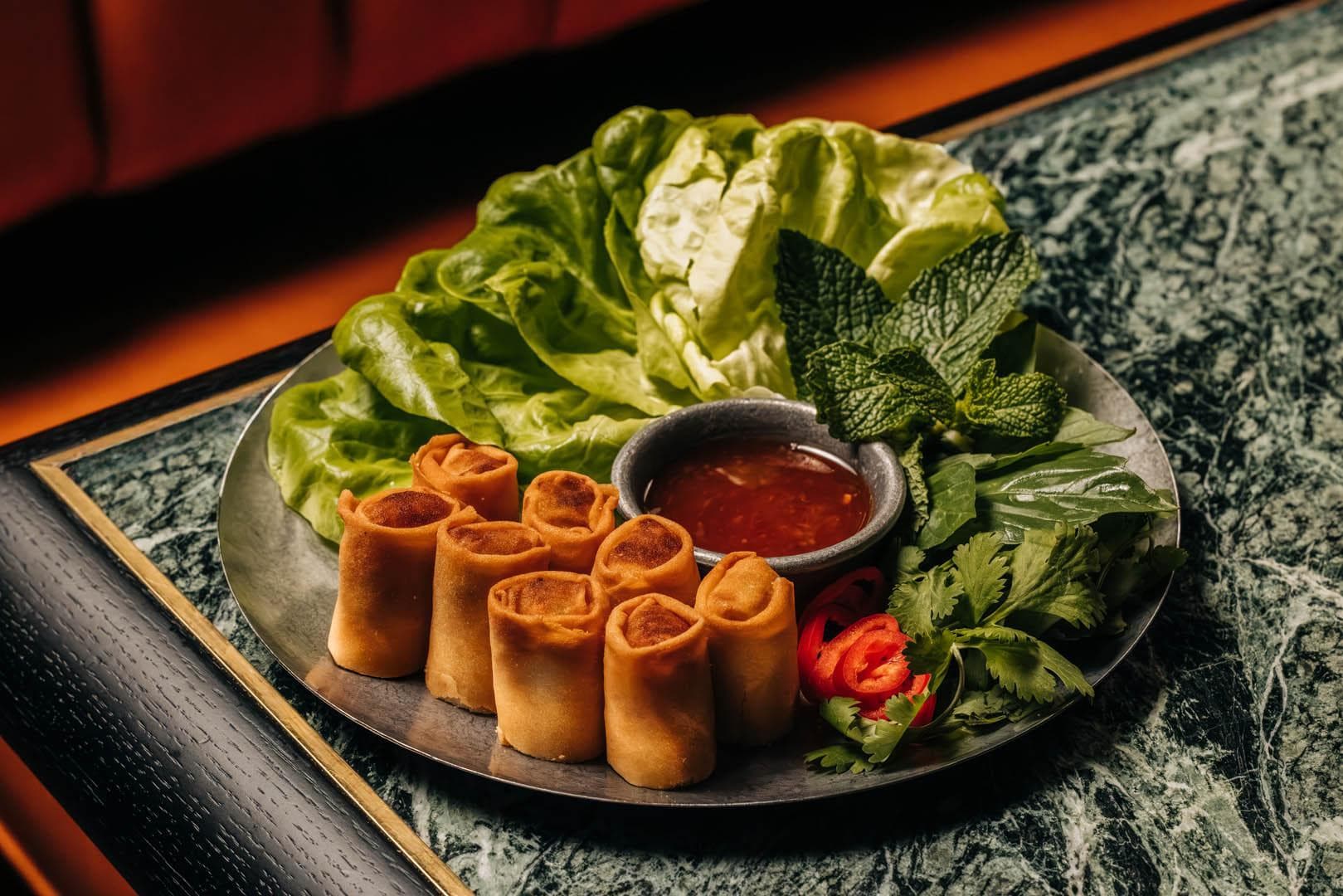 A plate of neatly arranged spring rolls with a small bowl of dipping sauce, surrounded by fresh lettuce, mint leaves, and sliced chili peppers.